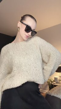 Camisola bege oversized de malha com mangas largas e corte solto, combinada com saia preta de cintura alta e óculos de sol grandes pretos.