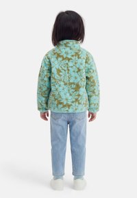 Dziecko o ciemnych włosach stoi tyłem, ubrane w niebiesko-zieloną polarową kurtkę w wzory, jasnoniebieskie dżinsy i białe trampki.