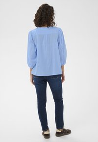 Blouse bleu clair, ample, avec des manches bouffantes et un détail froncé au dos, associée à un jean en denim foncé et des chaussures marron.