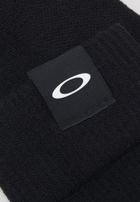 Schwarze Strickmütze mit strukturiertem Muster und einem rechteckigen schwarzen Etikett mit einem weißen Logo auf der Vorderseite.