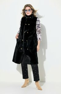 Gilet lungo in pelliccia sintetica nera con bottoni dorati, abbinato a un top fantasia e pantaloni neri decorati con accenti di perle bianche. Stivali alla caviglia beige completano l'outfit.