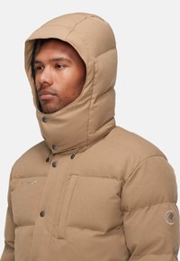 Giacca puffer isolante color sabbia con colletto alto e cappuccio, caratterizzata da bottoni a pressione scuri, una tasca sul petto e una toppa con logo sulla manica.