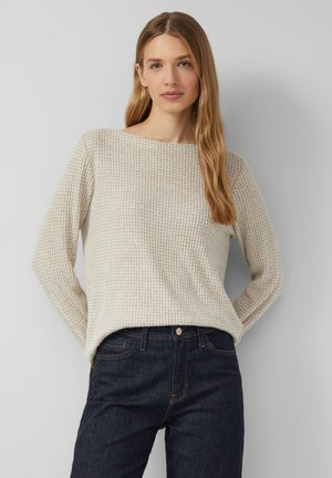 Donna con lunghi capelli biondi che indossa un maglione a maniche lunghe beige con un motivo e jeans blu scuro, in piedi di fronte a uno sfondo grigio semplice.