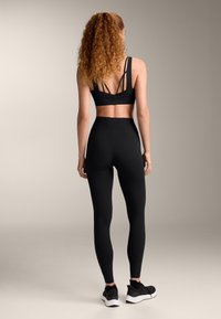 Musta urheiluliivi, jossa on useita ohuita hihnoja selässä ja korkeavyötäröiset mustat leggingsit. Mallilla on päällään mustat urheilukengät. Sileä pinta.