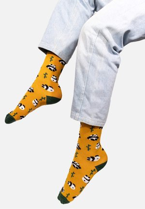 Żółte skarpetki w wzory pandy i kaktusa, z zielonymi akcentami na palcach i piętach, wykonane z mieszanki bawełny, długość do kostek, noszone z jasnoniebieskimi dżinsami.