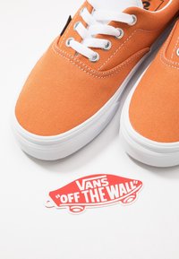 Baskets en toile orange avec lacets blancs, semelles en caoutchouc, œillets en métal et étiquette Vans "Off the Wall" en rouge et blanc.