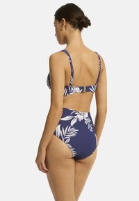 Model odwrócona, ubrana w granatowy bikini w białe kwiatowe wzory. Widoczne są eleganckie majtki wysokiego stanu oraz regulowane ramiączka.