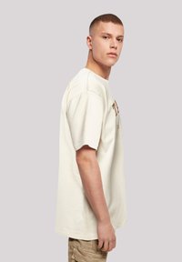 Light beige t-shirt with short sleeves, relaxed fit, and front print in brown. Fabric appears soft with a smooth texture.