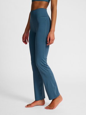 Frau trägt hoch taillierte, blaue eng anliegende Yogahosen und steht barfuß auf weißem Boden vor einem schlichten Hintergrund.