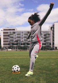 Completo sportivo grigio con accenti rosa, strisce nere e tessuto testurizzato. Il giocatore colpisce un pallone da calcio bianco e nero su un campo.