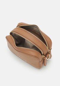 Hnědá kožená crossbody taška se dvěma otevřenými zlatými zipy, které odhalují prázdné vnitřní přihrádky na bílém pozadí.