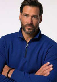 Blauer Zip-Kragen-Pullover aus einem weichen, strukturierten Stoff, mit einem Viertelreißverschluss und einer schlanken Passform. Das Modell hat die Arme verschränkt.