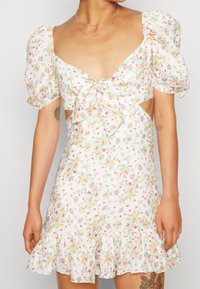 Femme portant une mini-robe blanche à imprimé floral avec manches bouffantes, nouée à l'avant, découpes latérales et ourlet volanté, dévoilant des tatouages sur le bras et la cuisse.