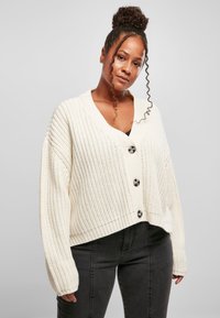 Chunky stickad ivorifärgad cardigan med V-ringning, tre stora svarta knappar och neddragna axlar. Ribbad nederkant och ärmslut, kort designer.