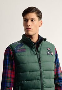 Chaleco acolchado verde con cierre de cremallera, con el logo del Oxford Polo Team y tres insignias. Lleva puesto sobre una camisa de cuadros en rojo, azul y verde.