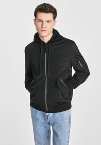 Czarna bluza z kapturem na zamek, ze sznurkiem do regulacji, bocznymi kieszeniami oraz kieszenią na rękawie zapinaną na zamek, wykonana z gładkiego materiału. Noszona z jasnoniebieskimi dżinsami.