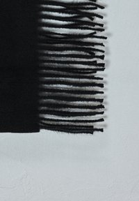 Črn volnen šal z dolgimi, enakomerno razporejenimi resicami na svetlo sivi teksturirani površini, ki prikazuje mehko tkanino in podrobnosti senc.