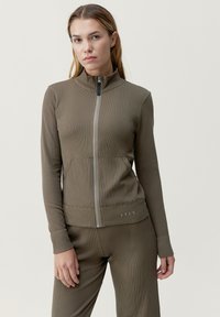 Giacca marrone con zip e colletto alto, texture a coste, vestibilità slim e tasche laterali. Presenta un logo discreto all'orlo e maniche lunghe.