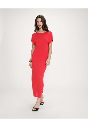 Femme en robe rouge longue jusqu'aux chevilles et talons ouverts noirs, debout devant un fond blanc uni.