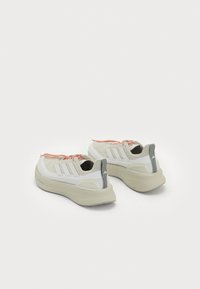 White athletic shoes with textured grey soles, orange laces, and three grey stripes on the side. Heel features vertical grey accent.