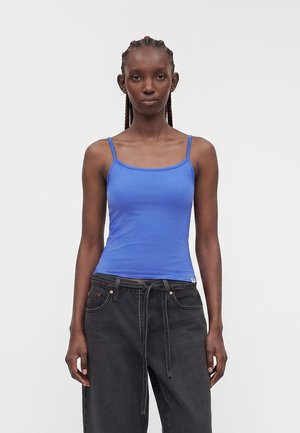 Kvinde med flettet hår iført en blå spaghetti-strop top og løstsiddende sorte jeans, stående mod en ensartet lys baggrund.