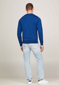 Blauer gestrickter Pullover mit Rundhalsausschnitt, geripptem Hals- und Ärmelabschlüssen, getragen mit hellblauen Jeans und weißen Sneakers, von hinten angesehen.