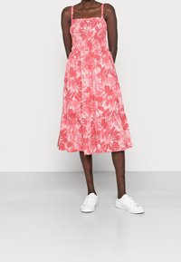 Robe mi-longue fleurie rose avec corsage élastique, fines bretelles et bas à volants. Portée avec des baskets blanches sur un fond uni.