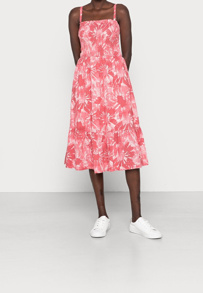 Robe mi-longue fleurie rose avec corsage élastique, fines bretelles et bas à volants. Portée avec des baskets blanches sur un fond uni.