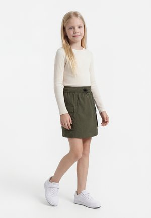 Ung jente med langt blondt hår iført en kremfarget ribbestrikket langermet topp, olivengrønn skjørt med lommer og hvite joggesko, står selvsikkert.