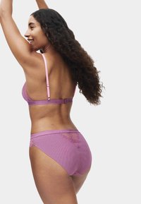 Kobieta z długimi kręconymi włosami, ubrana w różowy koronkowy biustonosz i dopasowane majtki, unosząca ręce i uśmiechająca się, widziana od tyłu na białym tle.