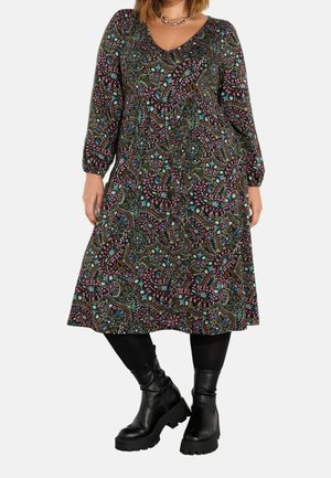 Donna che indossa un abito a maniche lunghe, lungo fino al ginocchio, con motivo paisley colorato, leggings neri, stivali neri robusti e una collana a catena d'argento.