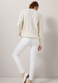 Pull en tricot torsadé crème à manches longues, associé à un jean blanc ajusté et des chaussures à talons, vu de dos sur un fond neutre.