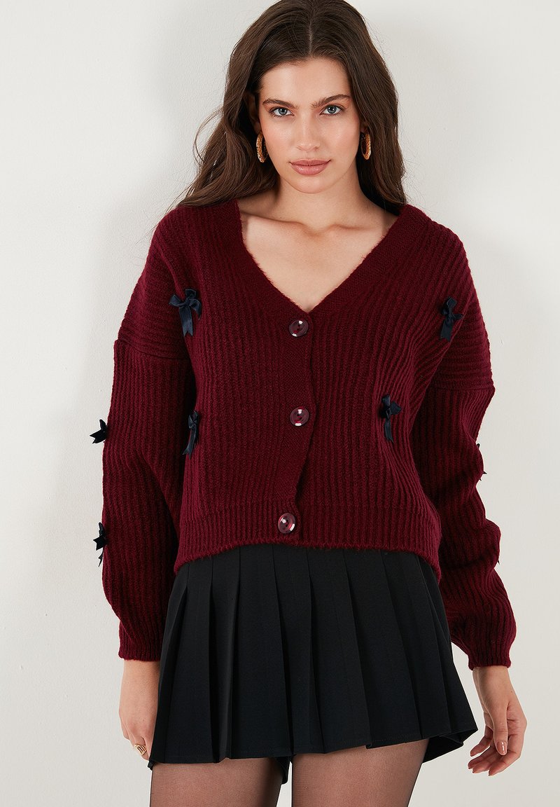 Burgundy stickad cardigan med V-hals, med stora knappar och marinblå banddetaljer på ärmarna. Texturerad, avslappnad passform.