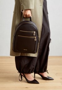 Mochila negra de cuero sintético con dos bolsillos frontales con cremallera y acentos en dorado. Forma redondeada y asa en la parte superior. Textura suave.
