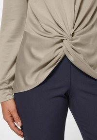 Beige Langarmoberteil mit einem Knoten-Detail an der Taille, kombiniert mit eng anliegenden dunkelblauen Hosen. Glatte Textur und subtiler Glanz sind offensichtlich.