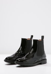 Botines Chelsea de charol negro brillante con paneles laterales elásticos, tiradores y detalles de brogue en la punta y los lados.