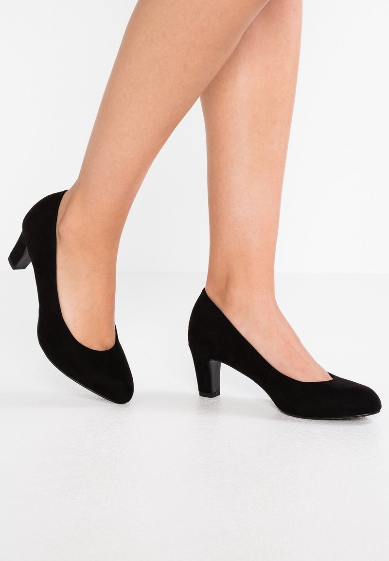 Pieds portant des escarpins noirs en daim à talons moyens et bouts arrondis, posés sur un sol blanc devant un fond blanc uni.