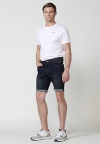 Camiseta de algodón blanca con logotipo negro, combinada con pantalones cortos de mezclilla oscura doblados en el dobladillo. Lleva unas zapatillas deportivas grises y blancas.