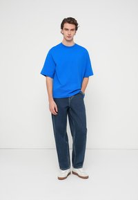 Μπλε κοντομάνικο T-shirt με άνετη εφαρμογή, συνδυασμένο με τζιν παντελόνι ευθείας γραμμής σε σκούρο ντένιμ και λευκά αθλητικά παπούτσια με καφέ σόλες.