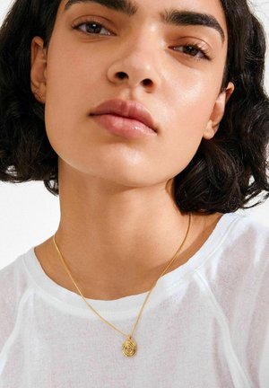 Zlata verižica z okroglim obeskom, ki ima spiralo, nošena na goli vratu, na svetli podlagi z belo majico.