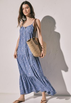 Femme en robe maxi bleue à motifs floraux portant un grand sac cabas tissé, debout pieds nus contre un mur clair texturé avec une ombre.