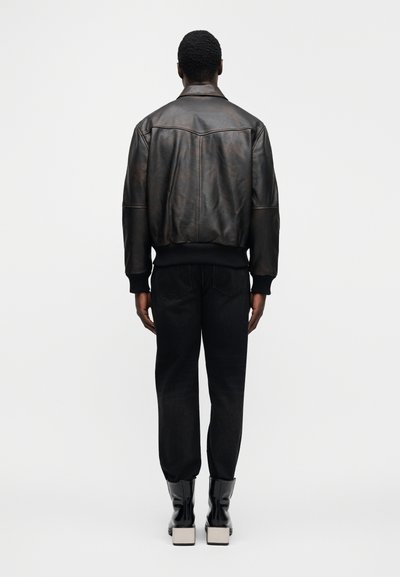 Καφέ δερμάτινο μπουφάν μπόμερ με ριμπ μανσέτες και μέση, σε συνδυασμό με σκούρο τζιν παντελόνι και γυαλιστερά μαύρα μποτάκια στο αστράγαλο.