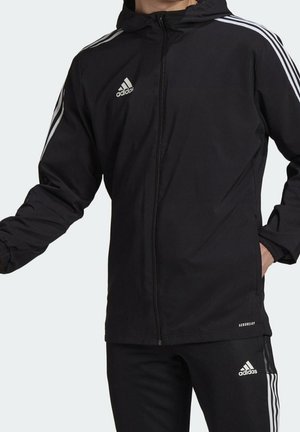 Personne portant une veste à capuche Adidas noire et un pantalon avec des bandes blanches et des logos, main droite dans la poche, bras gauche tendu.