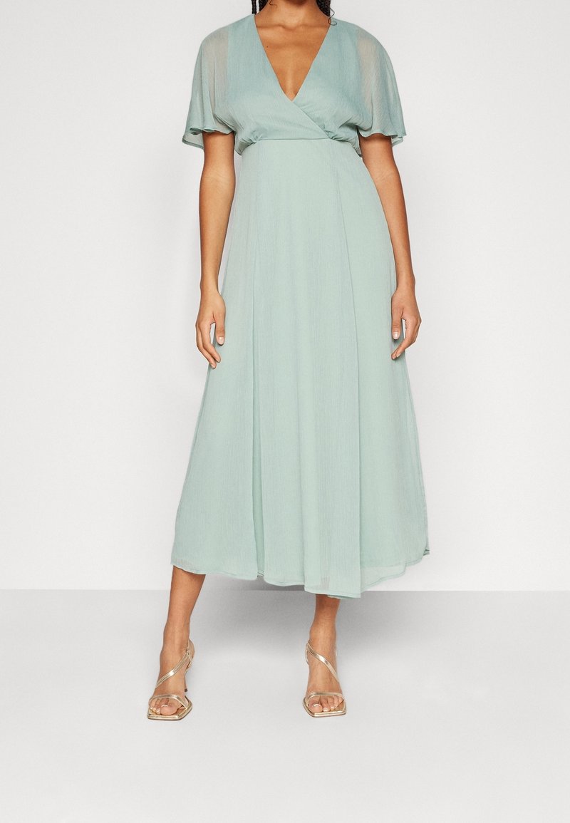 Femme portant une robe midi vert clair à manches courtes volantées et encolure en V, associée à des sandales à talons hauts dorées à brides.