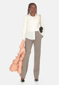 Cardigan beige e camicia bianca con bottoni, abbinati a pantaloni marrone chiaro. Tieni in mano un giubbotto trapuntato rosa brillante; una borsa nera completa l'outfit.