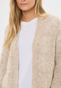Beżowy sweter z dzianiny z otwartym przodem, z teksturowanymi wzorami i subtelnymi metalicznymi akcentami. Nałożony na biały T-shirt. Lekki i przewiewny design.