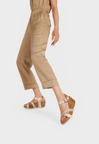 Sandali con zeppa e suola in sughero, con cinturini incrociati beige e una fibbia metallica. Il modello indossa pantaloni cropped beige.