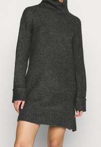 Mörkgrå, ribbstickad turtleneck klänning med långa ärmar och vikta manschetter, bärs av en person med bara ben.