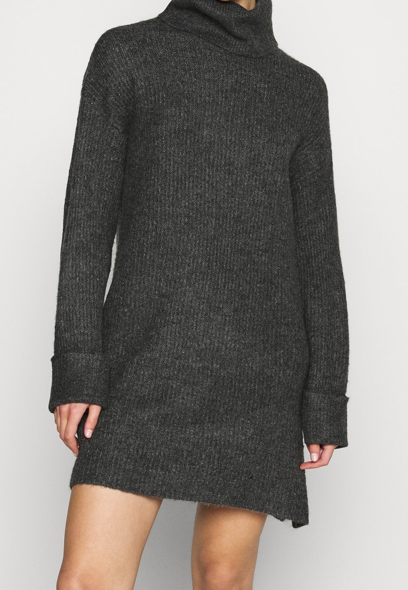 Mörkgrå, ribbstickad turtleneck klänning med långa ärmar och vikta manschetter, bärs av en person med bara ben.