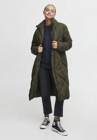 Lang, olivgrønn dunjakke med høy krage, som har en quiltet tekstur og en frontglidelås. Kombinert med mørke jeans og svarte joggesko.